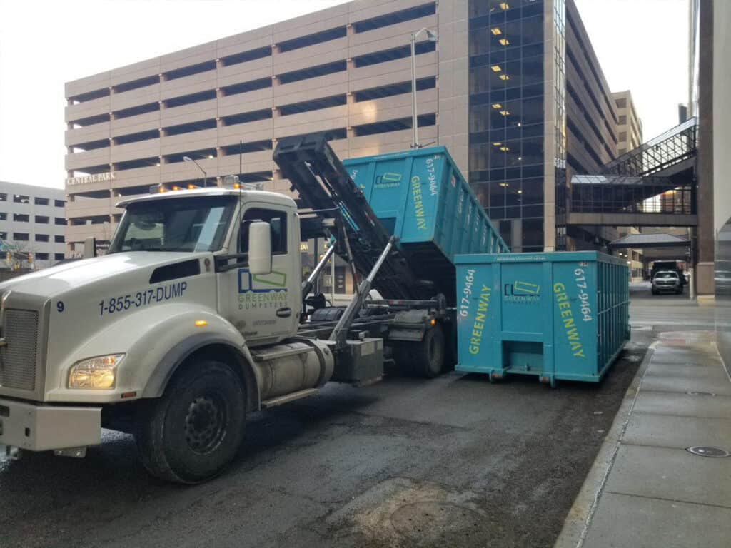 Dual Dumpsters in Action: Unloading and Placement Two dumpsters, one being unloaded next to the other in a scenic location